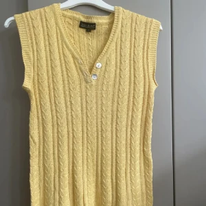 Gul stickad topp från Vero Moda - Snygg gul stickad topp från Vero Moda med kabelstickat mönster och v-ringning. Västen har två knappar framtill och är ärmlös, perfekt att styla över en skjorta eller t-shirt. Materialet är en mjuk mix av viskos och bomull.