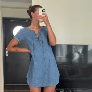 Blå kort jeansklänning från Lindex - Säljer en blå kort jeansklänning från Lindex med snörning framtill och korta ärmar. Klänningen har en avslappnad passform och är gjord i mjukt denimtyg, perfekt för varma dagar. Enkel och stilren design som är lätt att matcha.
