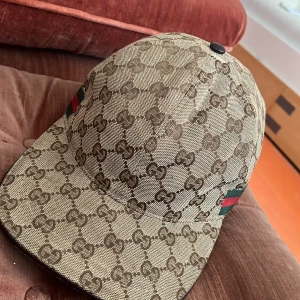 Gucci keps - Snygg keps från Gucci med klassiskt GG-monogram i brunt och beige. Kepsen har gröna och röda ränder på sidan och justerbar rem i mörkbrunt skinn baktill. Tillverkad i canvas och skinn, perfekt för dig som vill sticka ut med en lyxig accessoar.PRIS KAN DISKUTERAS 