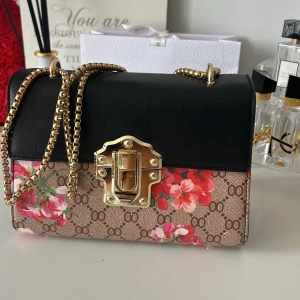  axelväska med blommigt mönster - Snygg axelväska med klassiskt GG-mönster och färgglada blommor. Väskan har svart lock i skinn, guldfärgad kedja och låsdetalj framtill. Insidan har dragkedjefack för småsaker. Perfekt för dig som vill sticka ut med en lyxig och trendig accessoar.
