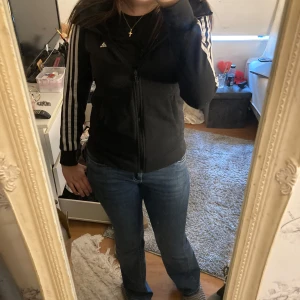 Svart Adidas hoodie med dragkedja - Oppss kom privat köp inte i Plick Svart hoodie från Adidas med klassiska vita ränder längs ärmarna och dragkedja framtill. Hoodien har huva, två fickor och är långärmad. Perfekt för en sportig look och enkel att matcha med jeans eller träningskläder.