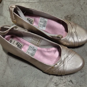 Ballerinaskor - Snygga beige ballerinaskor med kilklack och rund tå. Skorna har dekorativa remmar framtill med små guldfärgade nitar och en mjuk rosa innersula. Perfekta för dig som vill ha en klassisk men ändå trendig look.