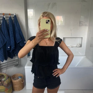 Svart plisserad blus med volanger - Supersnygg svart blus med plisserat tyg och dubbla volanger. Den har breda axelband och en rak, luftig passform som ger en cool och trendig look. Perfekt att styla med shorts eller jeans för en chic vibe.