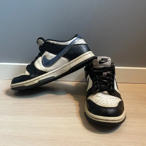 Nike Dunk Low svart/vit/blå 36.5 - Nike Dunk Low sneakers i svart, vitt och blått med klassisk swoosh i blått på sidorna. Skorna har snörning, perforerad tå och platt sula. Bekväm mesh på insidan och tydlig Nike-logga på häl och plös. Perfekt för dig som gillar retrovibbar och streetstyle.