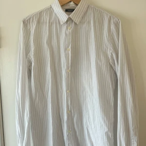 Vit randig skjorta från Filippa K - Klassisk vit skjorta från Filippa K med tunna grå ränder. Skjortan har lång ärm, knappar framtill och en stilren krage. Tillverkad i bomull för en skön och luftig känsla. Storlek är M. 