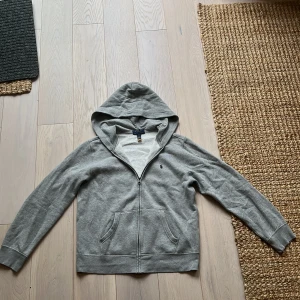 Grå hoodie från Polo Ralph Lauren - Klassisk grå hoodie från Polo Ralph Lauren med dragkedja framtill och två fickor. Tröjan har huva, långa ärmar och en liten broderad logga på bröstet. Tillverkad i mjuk bomullsmix som är skön att ha på sig hela dagen. tröjan är inpicip oanvänd och är storlek 14-16 år så för någon med längden cirka 165-175 cm. Hör av dig ifall du har några frågor eller funderingar 👍