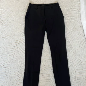 Svarta lågmidjade kostymbyxor från Ellos - Säljer ett par klassiska svarta kostymbyxor från Ellos med raka ben, hällor för bälte och diskreta fickor bak. Byxorna har dragkedja och knapp framtill och är tillverkade i ett mjukt material som känns bekvämt.