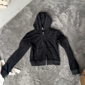 Svart velourjacka Juicy Couture - Svart velourjacka från Juicy Couture med huva och dragkedja framtill. Klassisk JC-logga broderad på bröstet och två fickor. Mjuk och skön med ribbade muddar vid ärmslut och midja. Perfekt för en chill och trendig look.