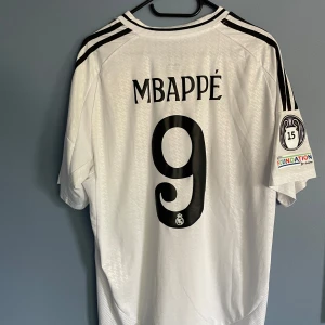 Real Madrid Mbappé 9 Adidas tröja - Säljer en vit Real Madrid fotbollströja med Mbappé och nummer 9 på ryggen. Tröjan har svarta detaljer, broderat klubbmärke och Adidas-logga, samt Champions League-märke på ärmen. Tillverkad i lätt och ventilerande material, perfekt för match eller träning.