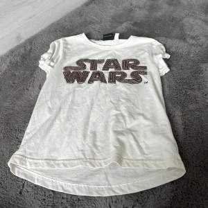 Vit Star Wars t-shirt med pärldetaljer - Vit t-shirt från Star Wars med coolt pärlbroderat logotyptryck på bröstet. Klassisk rund hals, korta ärmar med rynkade detaljer och något längre bakstycke. Perfekt för alla Star Wars-fans som vill sticka ut.