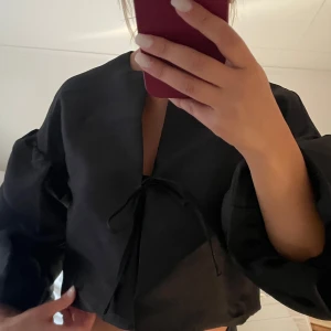 Svart topp med knyt från Gina  - Säljer en svart topp/ blus med knyt framtill och vida ärmar. Blusen är croppad och har en stilren, minimalistisk look. Perfekt att styla med höga jeans eller kjol för en trendig vibe. Från Gina! Passar xs och s. 