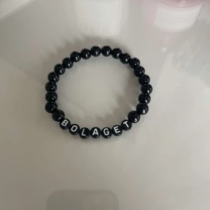 Svart pärlarmband med text - Snyggt svart pärlarmband med bokstäver som bildar ordet 'BOLAGET'. Perfekt accessoar för en personlig touch. Elastiskt band för enkel passform.