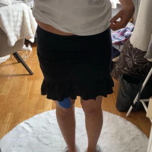 Svart kjol med volang - Snygg svart kortkjol med volangdetalj längst ner. Perfekt för en stilren look. Kjolen har en tajt midja men den är också strechig. 