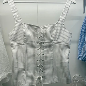 Vit randig korsett med snörning - Snygg vit korsett med svarta ränder och snörning framtill. Den har breda axelband och en stilren design som passar perfekt för en trendig look.