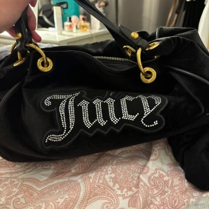 Svart axelväska från Juicy Couture - Snygg svart axelväska från Juicy Couture med glittrande detaljer och guldfärgade metalldelar. Väskan har en dragkedja och ett inre fack med Juicy Couture-mönster. Perfekt för att ge en touch av glamour till din outfit.