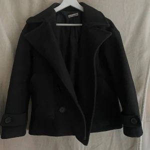 Svart kappa från Design by Si - Elegant svart kappa från Design by Si med breda slag och knappar framtill. Perfekt för kyligare dagar med sin klassiska design och stilrena look.