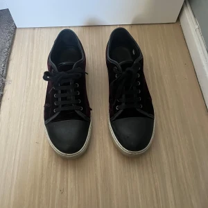 Vinröda lanvin skor - Snygga sneakers från Lanvin i svart och vinröd sammet. Skorna har en klassisk snörning och en robust gummisula. Perfekta för en stilren look med en touch av lyx. Lite crease skador och lite skada där bak annars lugnt. Storlek 43-44