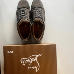 Grå sneakers från Christian Louboutin stolek 44 - Snygga grå sneakers från Christian Louboutin med den ikoniska röda sulan. Skorna har snörning och är tillverkade i mocka, vilket ger en lyxig känsla. Perfekta för dig som vill ha en stilren och exklusiv look.