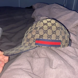 Beige keps från Gucci - Snygg beige keps från Gucci med det ikoniska GG-mönstret. Kepsen har en justerbar rem baktill i blått läder och en röd-blå randdetalj på sidan. Perfekt för att ge din outfit en lyxig touch.