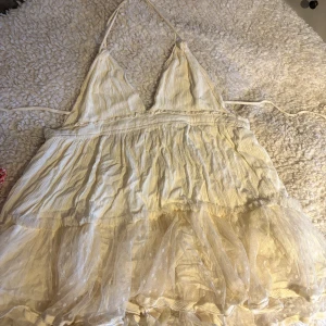 Vit klänning med volangdetaljer - Charmig vit klänning med smala axelband och volangdetaljer. Klänningen har en omlott-design och en lätt, luftig känsla med prickigt mönster på volangerna. Perfekt för varma sommardagar. Öppen i ryggen. 