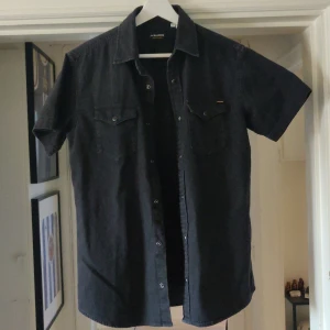Svart kortärmad skjorta - Säljer en stilren svart kortärmad jeans skjorta. Skjortan har knappar framtill och två bröstfickor med lock. Perfekt för en avslappnad look. Använd en gång hemma men satt inte så bra och etiketten va bort tagen redan