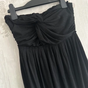 Ny jättesöt tubtoppklänning - Elegant svart axelbandslös klänning med en knutdetalj framtill. Klänningen har en plisserad överdel och ett flödande kjolparti som ger en fin siluett. Perfekt för en stilren look.