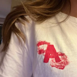 Vit t-shirt med rött tryck - Säljer en vit t-shirt med ett stort rött tryck på baksidan och ett lite mindre fram. Trycket har en konstnärlig stil med ett stort 'A' på framsidan. Perfekt för en avslappnad look!