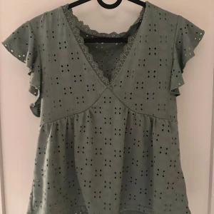 Grön blus med spetsdetaljer - Charmig grön blus med korta ärmar och spetsdetaljer. Blusen har ett vackert hålmönster och en v-ringad design. Perfekt för en stilfull och avslappnad look.