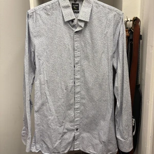 Vit och blå mönstrad skjorta från Olymp - Säljer en stilren vit med blåa blommor skjorta från Olymp. Skjortan har en super slim passform och långa ärmar, perfekt för en elegant look. Den är tillverkad i ett bekvämt material och har klassiska knappar framtill.