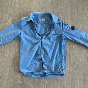 Blå overshirt från C.P. Company - Säljer en snygg blå overshirt från C.P. Company med dragkedja och en cool detalj på ärmen. Perfekt för en stilren look. 