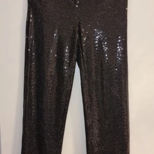 Svarta glittriga byxor  st S  helt nya - Snygga svarta byxor med glittrig yta som verkligen fångar ljuset. Perfekta för en kväll ute eller när du vill sticka ut lite extra. De har en rak passform och är tillverkade i ett syntetmaterial som ger en bekväm känsla.