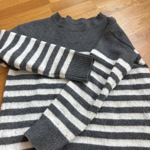 Grå och vit randig stickad tröja - Säljer en mysig grå och vit randig stickad tröja med långa ärmar. Perfekt för kyliga dagar med sin ribbade hals och muddar. En klassisk och stilren design som passar till mycket.