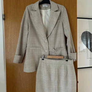 Beige rutig kavaj och kjol, AFJ kollektionen från Nakd - Snyggt set med kavaj och kjol i beige och vitt rutigt mönster från Nakd AFJ kollektionen.  Kavajen har två knappar och klassisk krage, medan kjolen är knälång och stilren. Perfekt för en chic och professionell look. Limited Edition utgåva! Kavaj använd 1 gång, kjolen är aldrig använd! Kavajen storlek 36 och kjolen storlek 38