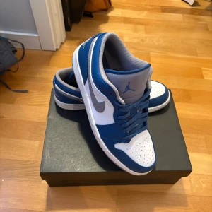 Nike Air Jordan 1 Low i blått och vitt - Säljer ett par Nike Air Jordan 1 Low i en snygg blå och vit färgkombination. Skorna har den klassiska Air Jordan-loggan på hälen och en vit sula. Perfekta för dig som vill ha en stilren och sportig look.