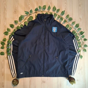 Mörkblå OS 2004 vindjacka från Adidas - Säljer en sällsynt mörkblå vindjacka från Adidas med broderad logga och OS Athens 2004-märke på bröstet. Jackan har dragkedja framtill och två fickor med dragkedjor. Den har långa ärmar med vita ränder längs sidorna och elastiska ärmslut.