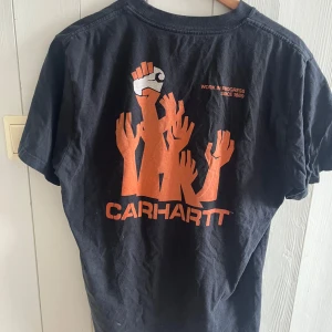 Svart t-shirt från Carhartt - Säljer en svart t-shirt från Carhartt med ett coolt tryck av uppsträckta händer i orange på baksidan och en liten knuten näve på framsidan. Perfekt för en avslappnad stil.