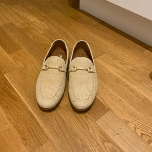 Beige loafers med gulddetaljer.  - Snygga beige loafers med elegant gulddetalj på ovansidan. Perfekta för en stilren look. Tillverkade i ett texturerat material som ger en unik känsla. 