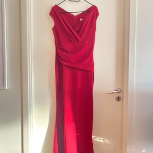 Röd långklänning med omlott - Elegant röd långklänning med omlott-design och offshoulder-stil. Perfekt för festliga tillfällen med sin smickrande passform och vackra drapering. Klänningen har en djup v-ringning och en subtil slits för extra rörelsefrihet.