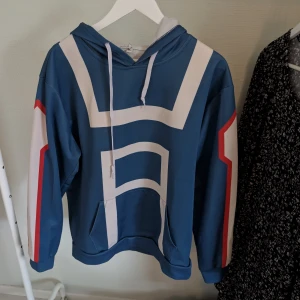 My Hero Academya merchandise  - Säljer en My Hero Academya blå hoodie med vita och röda detaljer. Den har en stor huva och en klassisk magficka framtill. Perfekt för en avslappnad stil.