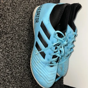 Blå Adidas Predator fotbollsskor - Snygga blå Adidas Predator fotbollsskor med svarta detaljer och snörning. Perfekta för inomhusspel med en slitstark sula. Skorna har en modern design med klassiska tre ränder på sidan.