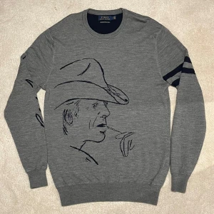 Ralph Lauren - Merinoull - Snygg grå tröja från Polo Ralph Lauren med ett stort cowboymotiv på framsidan. Tröjan har långa ärmar och mörkblå detaljer, inklusive två ränder på ena ärmen och siffran '096' på baksidan. Dessutom i merinoull vilket gör den borde skön och väldigt unik🙌 vid minsta fråga eller fundering hör av er🤝