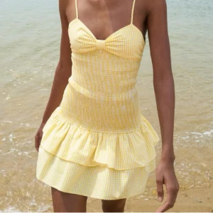 Gul rutig kortklänning med rosett Zara - Supersöt gul och vit rutig kortklänning med smala axelband, volangkjol och stor rosett i ryggen. Klänningen har smockad midja och omlottkänsla baktill. Perfekt för varma dagar och ger en riktigt somrig vibe.