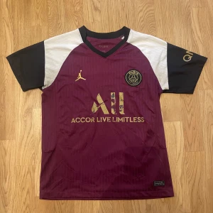 PSG vinröd matchtröja Nike Jordan 2018 - Snygg PSG fotbollströja i vinrött med svarta och vita detaljer. Tröjan har korta ärmar, guldiga loggor och Paris Saint-Germain-emblem på bröstet. Tillverkad i lätt och ventilerande material, perfekt för träning eller match. Jordan-logga på framsidan och Nike-märke nertill. Årtal 2018