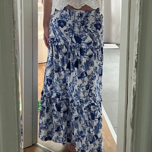 Blåvit blommig långkjol med snörning - Supersöt långkjol i vitt med blått blommönster och volanger. Kjolen har snörning med tofsar i midjan och är gjord i ett lätt, flowy material som känns perfekt för varma dagar. Passar dig som gillar romantisk och bohemisk stil.