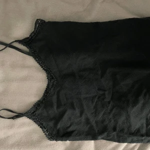 Svart linnetopp med spetskant - Snygg svart linnetopp med tunna axelband och dekorativ spetskant längs urringningen. Perfekt att styla med jeans eller kjol för en trendig look. Enkel och stilren design som passar till många outfits.