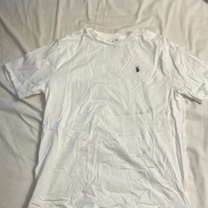 Vit t-shirt från Polo Ralph Lauren - Klassisk vit t-shirt från Polo Ralph Lauren med korta ärmar och rund hals. Ikoniska lilla broderade loggan på bröstet. Skön och mjuk bomull, perfekt till jeans eller shorts. Enkel och stilren design som alltid funkar.