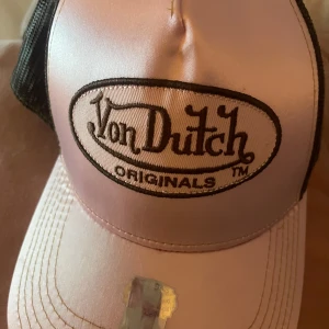 Rosa Von Dutch keps - Snygg rosa truckerkeps från Von Dutch med svart broderad logga framtill. Kepsen har svart mesh baktill för extra ventilation och klassisk böjd skärm. Insidan är dekorerad med Von Dutch-tryck och coola detaljer. Aldrig använd 