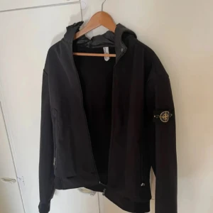 Svart vindjacka från Stone Island - Svart vindjacka från Stone Island med ikonisk patch på vänster ärm. Jackan har huva, dragkedja framtill och praktiska fickor med dragkedja. Perfekt för dig som gillar stilren och sportig look. Materialet är troligtvis polyester och jackan har en normal passform. Perfekt nu till sommaren