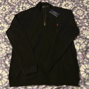 Svart half zip tröja Polo Ralph Lauren - Svart långärmad tröja från Polo Ralph Lauren med half zip-dragkedja och klassisk röd broderad logga på bröstet. Tröjan har hög krage och är tillverkad i mjuk bomull, perfekt för en clean och stilren look.