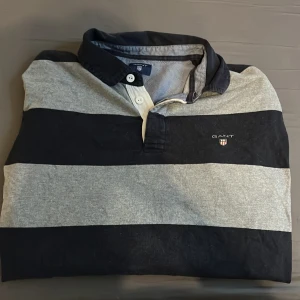 Randig pikétröja från GANT i svart och grå - Snygg långärmad pikétröja från GANT i storlek L. Tröjan har breda blåa och grå ränder, klassisk krage och knäppning framtill. GANT-logga broderad på bröstet. Perfekt för en avslappnad men stilren look. Hör av vid minsta lilla fråga 